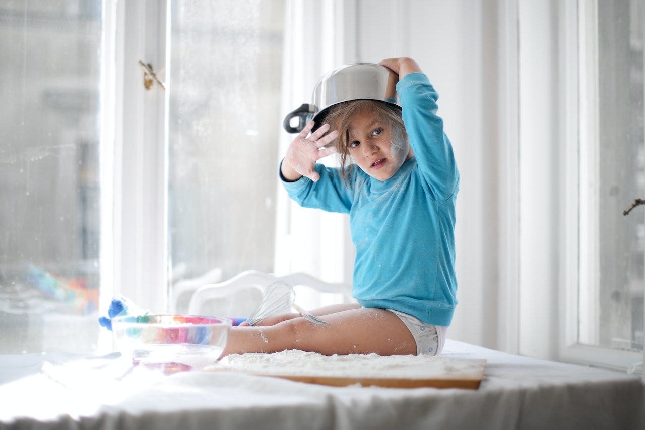 A young child sitting on a table with a bowl on her head
Description automatically generated with medium confidence