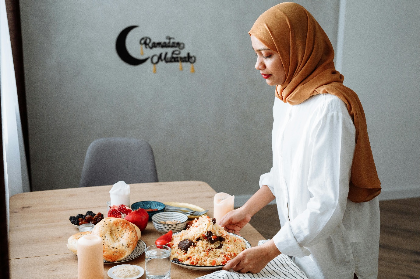 A person in a white shirt and brown head scarf holding a plate of food Description automatically generated